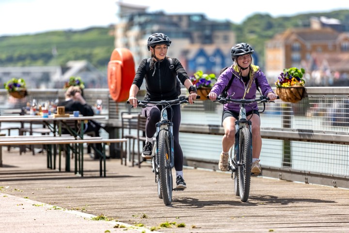 Two people cycling on a boardwalk near a waterfront, with flowers and buildings in the background.
