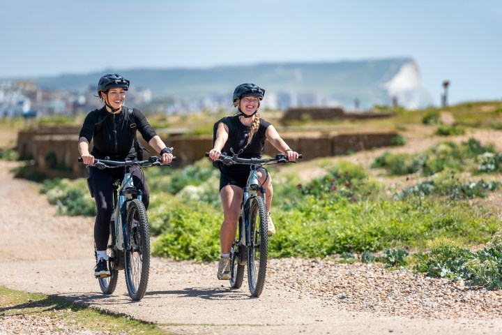 Two people biking on a path near the coast under a clear sky.