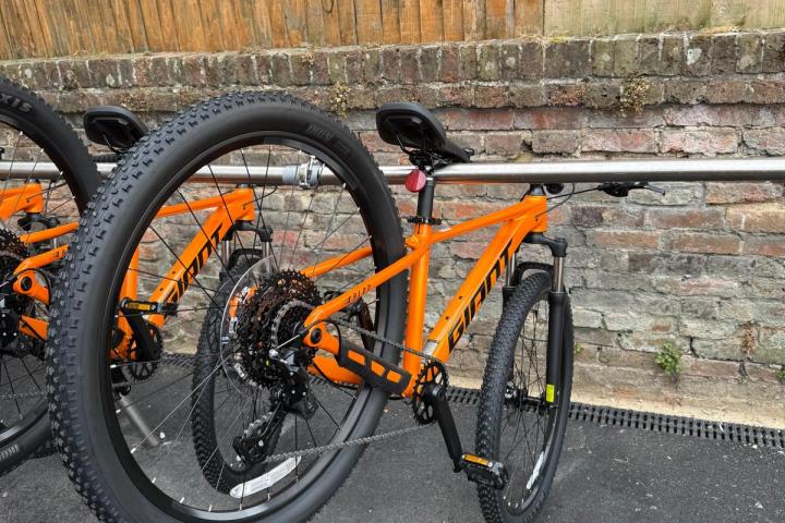 Orange mountain bike secured to a bike rack near a brick wall and wooden fence.